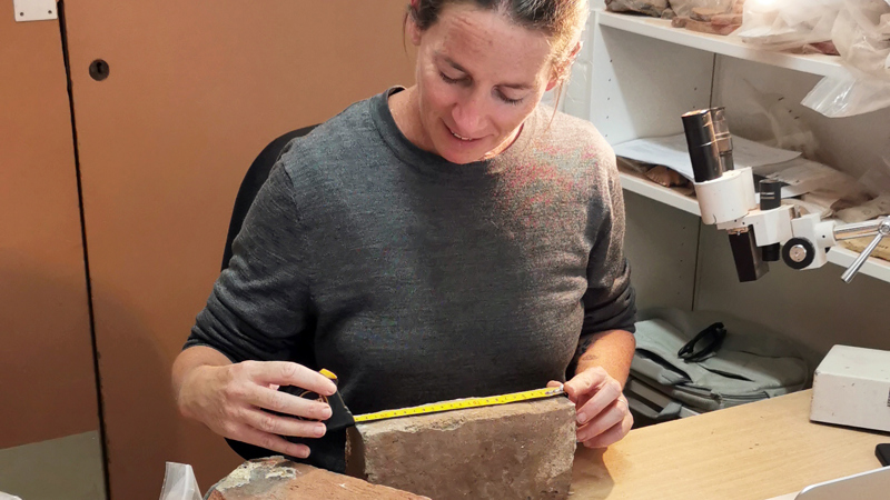 an archaeologist in a lab measures a brick with a yellow tape measure. Around her on the desk are other bricks, a small pickaxe for removing brick fragments, and a microscope.